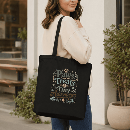 Woman carrying a black Paws, Treats & Tiny Adventures tote bag in a cozy lifestyle setting.